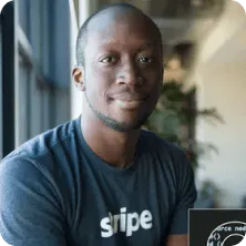 Man wearing a dark blue Stripe t-shirt sitting indoors near a window with a subtle smile.
